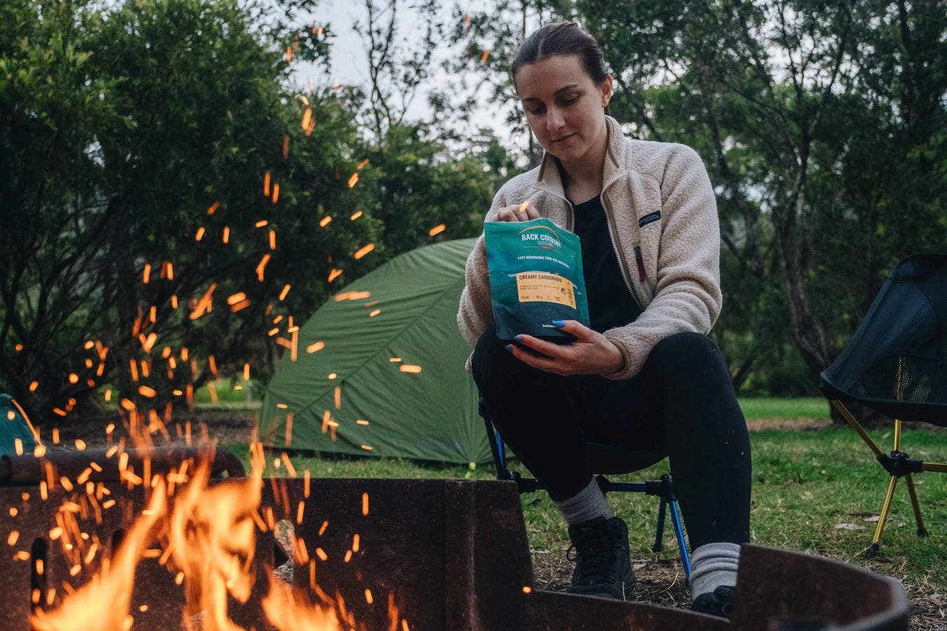 Person holding a package by a campfire with tents in the background